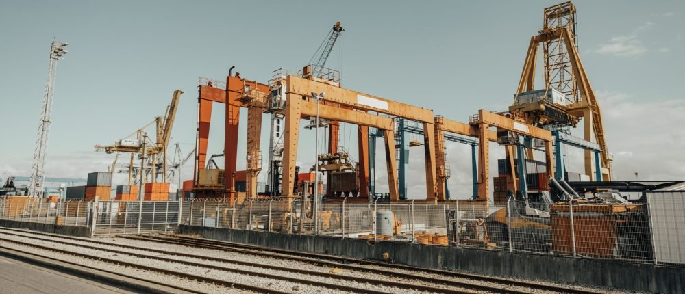 Large industrial port with multiple gantry cranes, shipping containers, and railway tracks. The heavy-duty structures, painted in yellow and orange, dominate the logistics hub under a clear sky